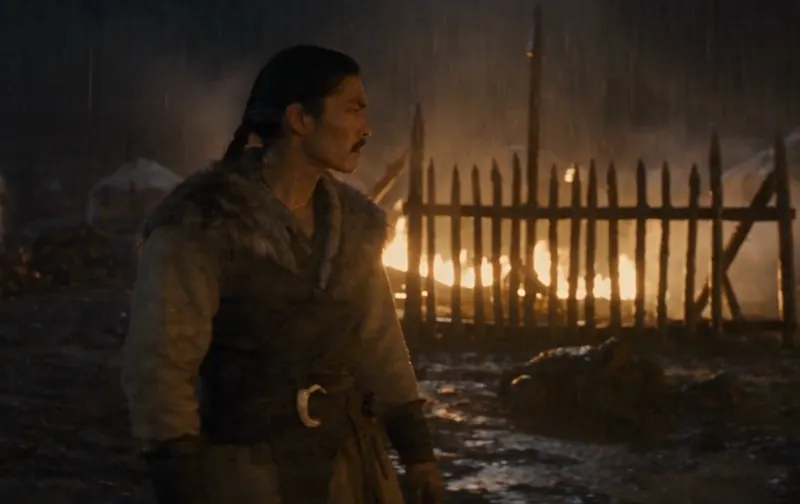 Still: Ashina in ruins. A man stands in the rain before a burning palisade.